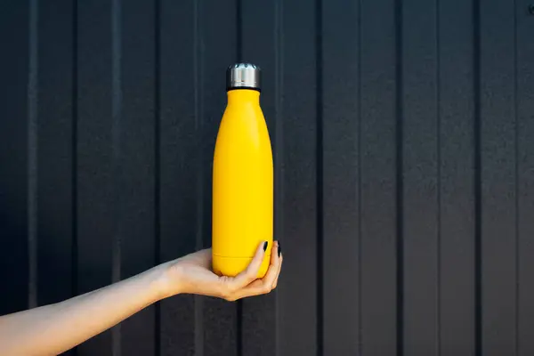 ブラックカラーのラインで金属の背景に黄色の鋼熱水ボトルを保持している女性の手のクローズアップ — ストック写真