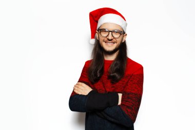 Kırmızı kazaklı, kolları çapraz Noel Baba şapkası ve gözlük takan gülümseyen genç adamın stüdyo portresi. Beyaz arkaplanda izole.