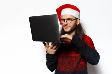 Kırmızı kazaklı, elinde dizüstü bilgisayarla Noel Baba şapkası ve gözlük takan genç bir adamın stüdyo portresi. Beyaz arkaplanda izole.