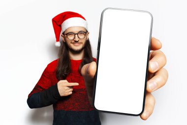 Stüdyo portresi, kırmızı kazaklı, ekranda boş olan ve Noel Baba şapkası ve gözlük takan büyük bir akıllı telefon tutan genç adam. Beyaz arkaplanda izole.