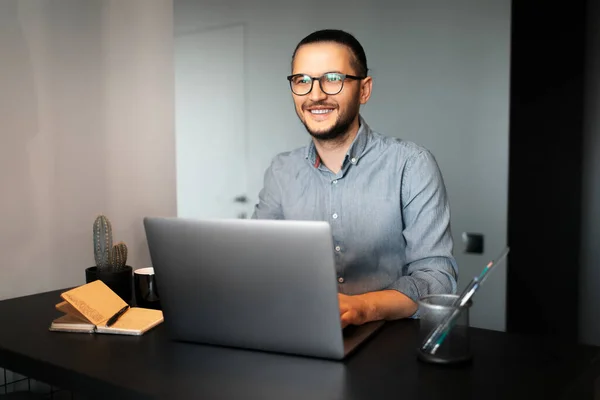 Portrait of young happy man working home at laptop, wearing eyeglasses and shirt, sitting at desk, background of grey wall.