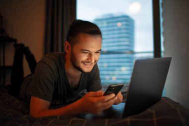 Akıllı telefon ve dizüstü bilgisayar kullanarak yatağa uzanan gülümseyen genç adam. Panoramik pencerenin arkaplanı.