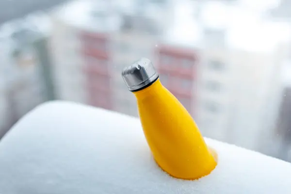 Steel thermo bottle of yellow color in snow. Close-up view. Reusable thermos.