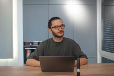 Laptop 'ta mutfakta çalışan düşünceli bir gencin portresi. İş konsepti. Gözlük ve kazak takıyor..