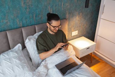 Telefonla yazı yazan genç bir adamın portresi, sabahları laptopta yatarken. İş adamları. Gözlük takıyordu. Mavi duvarın arka planı. Üst görünüm.