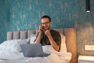 Genç gülümseyen bir adamın portresi. Sabah dizüstü bilgisayarla yatakta yatarken telefonla konuşuyor. İş adamları. Gözlük takıyordu. Mavi duvarın arkaplanı.