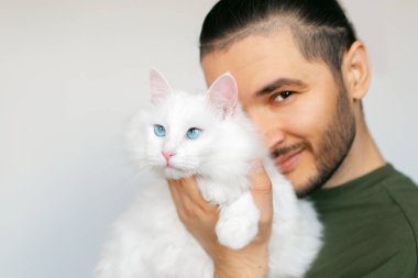 Beyaz arka planda mavi gözlü, beyaz tüylü kediyi kucaklayan gülümseyen genç adamın portresi..