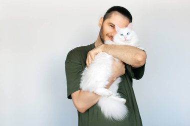 Beyaz arka planda mavi gözlü, beyaz tüylü kediyi kucaklayan gülümseyen genç adamın portresi..