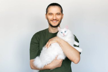 Beyaz arka planda mavi gözlü, beyaz tüylü kediyi kucaklayan gülümseyen genç adamın portresi..