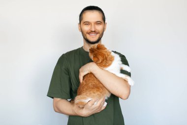 Beyaz arka planda pofuduk turuncu kediyi kucaklayan gülümseyen genç adamın portresi.