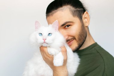 Beyaz arka planda mavi gözlü, beyaz tüylü kediye sarılan genç adamın portresi..