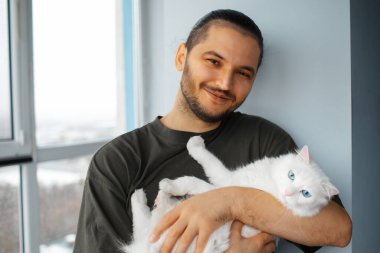 Mavi gözlü beyaz tüylü kediye sarılan mutlu genç adamın portresi..