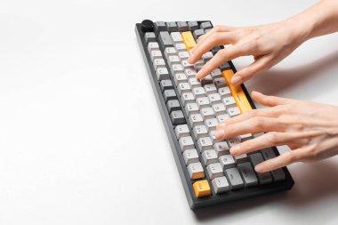 Beyaz arkaplanda, gri ve sarı anahtar kapaklı mekanik klavyede yazan kadın elleri.