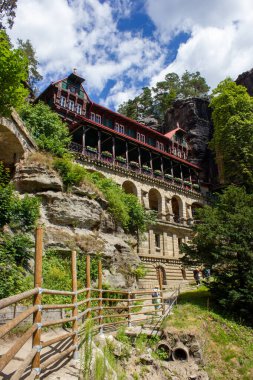 Mavi gökyüzü altında yeşil ağaçları olan kayalık dağlar arasında tarihi mimari yapı