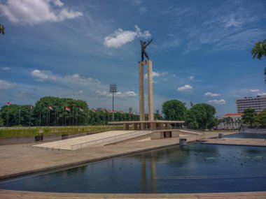 Ortasında gölet olan bir şehir parkı ve tepesinde ağaçlar ve parlak bulutlarla çevrili ikonik bir heykel olan uzun bir anıt.