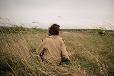 Uzun otların arasında otururken trençkot giyerek doğada poz veren manken.