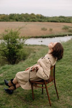 Açık havada huzurlu bir anın tadını çıkaran manken, gösterişli bej renkli bir trençkot ve botla sakin doğal bir arka plana karşı koyuyor.