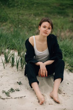 Kum tepeciğinde çıplak ayakla oturan manken, siyah takım elbise ve beyaz bluz giyiyor, açık havada fotoşop yaparken çarpıcı poz veriyor.