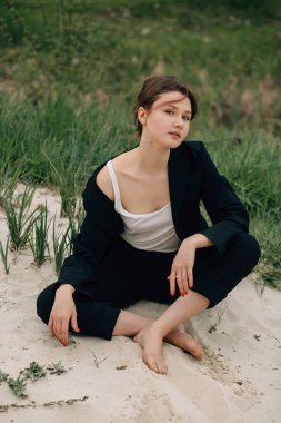 Kum tepeciğinde çıplak ayakla poz veren manken siyah takım elbise ve beyaz bluz giyiyor.