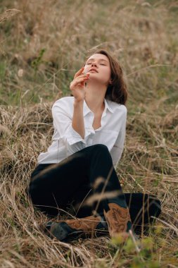 Moda mankeni doğada huzurlu bir anın tadını çıkarıyor, şık kovboy çizmeleri ve beyaz bir gömlek giyiyor.