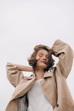 Trençkot giyen, gözleri kapalı dinlenen, elleri saçlarında, doğanın ve temiz havanın tadını çıkaran bir model.