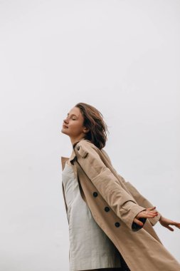 Bej renkli trençkot giyen manken, geniş kolları ve kapalı gözleriyle doğanın sükunetini kucaklıyor.