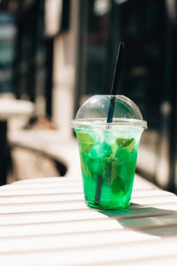 Parlak yeşil ferahlatıcı buzlu naneli içecek ve dışarıdaki beyaz bir masada siyah bir pipet oturuyor.