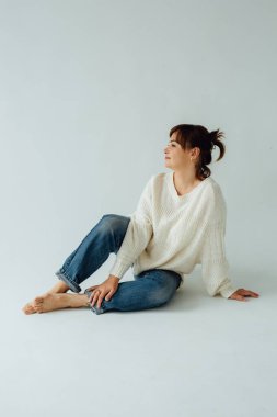 40 'lı yaşlarda, kot pantolon ve beyaz kazak giyen, yerde oturan ve yan tarafa bakan esmer bir kadının stüdyo portresi.