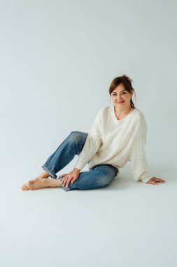 40 'larında, kot pantolon ve beyaz süveter giyen, beyaz bir arka planda rahat bir şekilde yerde oturan neşeli esmer bir kadının stüdyo portresi.