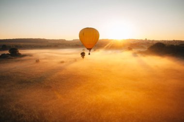 Sarı sıcak hava balonu sisli bir arazide uçuyor nefes kesen bir gün doğumunda büyülü bir atmosfer yaratıyor.