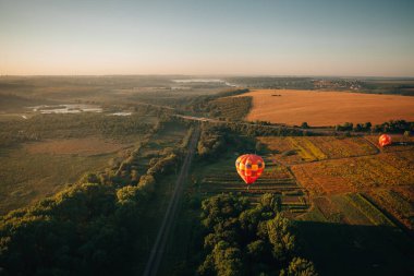 Renkli sıcak hava balonları gün doğumunda, resmedilmeye değer kırsal bir manzaranın üzerinde yavaşça süzülüyor, sakin ve nefes kesici bir manzara yaratıyor.