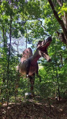 Bir dinozorun gerçek boyutlardaki heykeli kısmen yemyeşil yeşilliklerle kaplı güneşli bir ormanda saklanarak antik zamanların ve vahşi hayattaki maceraların anılarını canlandırıyor..