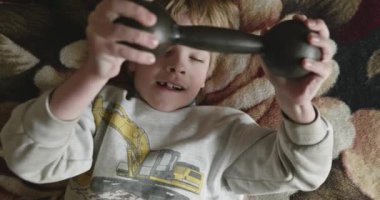 Çocuk evde dambıllarla egzersiz yapıyor. Çocuk, yatakta uzanarak egzersiz yapıyor. 4K Yavaş çekim 60 fps 10-bit