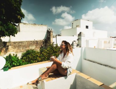 Bir kadın terasında kahve içerek rahatlar ve manzaraya bakar.