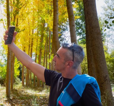 Bir adam ağaçların arasında cep telefonuyla selfie çekerken ormanda spor yapıyor.