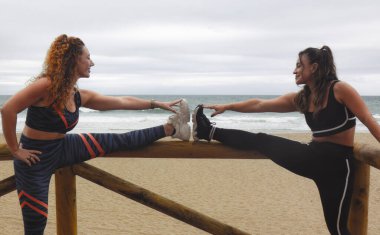 İki genç kadın sahilde esneme egzersizi yapıyor.