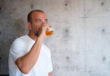 Beyaz tişörtlü bir adam evde kutlama yaparken bira içiyor.