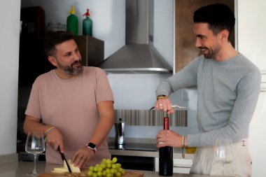 two lgtbi couple boys in the kitchen of their house preparing an appetizer, one of them opens a bottle of wine and the other cuts cheese clipart