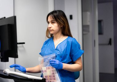 Laboratuvar teknisyeni mavi önlük ve eldiven giyiyor bilgisayar kullanıyor ve plastik bir çantada tıbbi örnekler tutuyor.