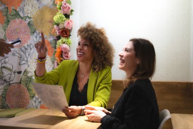 İki kadın yemek sipariş ediyor, biri seçim yapmak için parmağını kaldırıyor, bir restoranda çiçekli bir masada otururken.