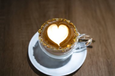 Pistachio latte sanatı kahvenin yüzeyinde bir kalp şekli yaratıyor, ahşap bir masada cam bir bardakta servis ediliyor.