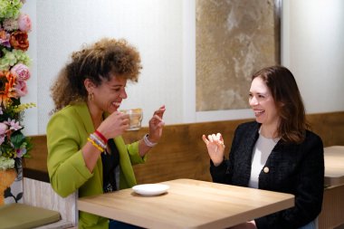 Kahve molası veren iki neşeli iş kadını bir kafede konuşurken gülüp el kol hareketi yapıyorlar.