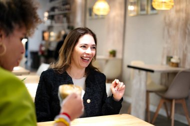 İki iş kadını kahve molası veriyor, modern bir kafede gülüyor ve sohbet ediyorlar.