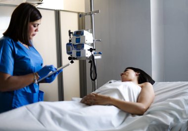 Hemşire dijital tablet kullanıyor. Kadın hastaların hastane yatağında serumla dinlendiklerini not alıyor.