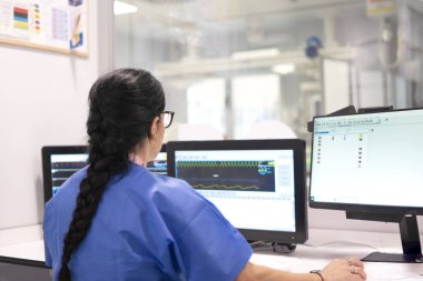 Hastane kontrol odasındaki bilgisayar ekranlarında görülen tıbbi verileri ve hastanın hayati belirtilerini analiz eden doktor.