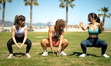 Parkta çömelme egzersizleri yapan üç bayan sporcu fiziksel aktivitenin faydalarından yararlanıyor.