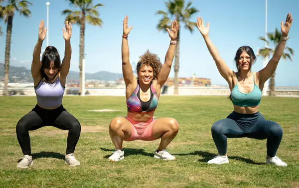 Üç kadın fitness eğitmeni, şehir parkındaki bir antrenman sırasında kollarını kaldırarak çömelme hareketleri yapıyorlar.