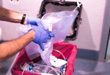Eldiven giyen hemşire hastane odasındaki biyolojik tehlike konteynırına kullanılmış plastik poşet atıyor.