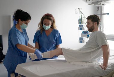 İki hemşire hastane odasında erkek bir hastanın bacağını bandajlıyor.