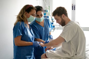 Maske ve eldiven takan iki hemşire bir hastayı hastane yatağında teselli ediyor, elini tutuyor ve zor zamanlarında ona destek oluyor.